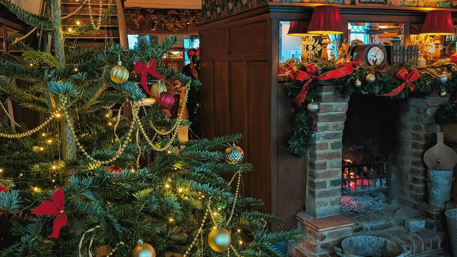 A Christmas tree with gold baubles and fairy lights beside a lit brick fireplace hung with garland and red bows at The Pullman, Folkestone