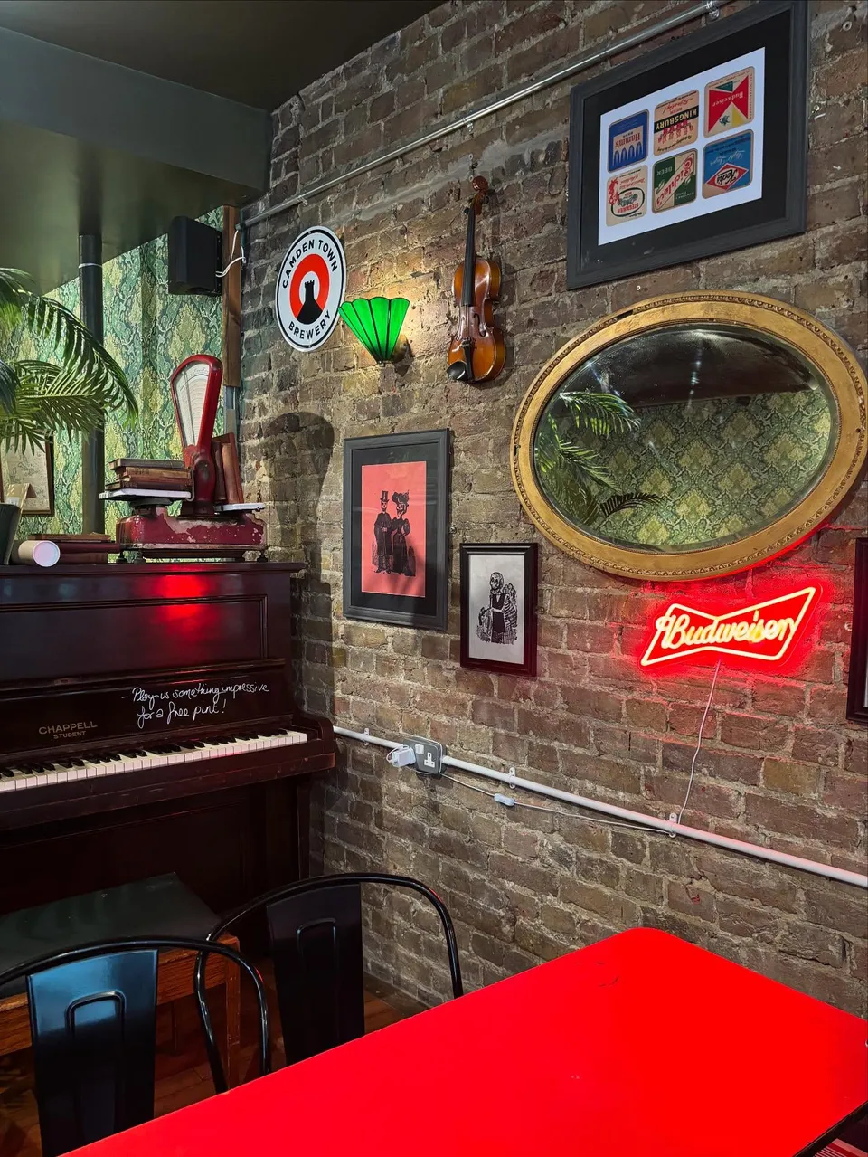 The piano corner at Cat & Fiddle: an upright piano under a fiddle and framed prints, beside a red Budweiser neon on bare brick