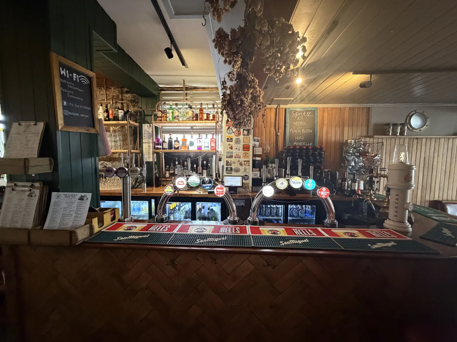 The bar at The Ship Inn in Sandgate, with green panelling, hanging hops and rows of beer taps